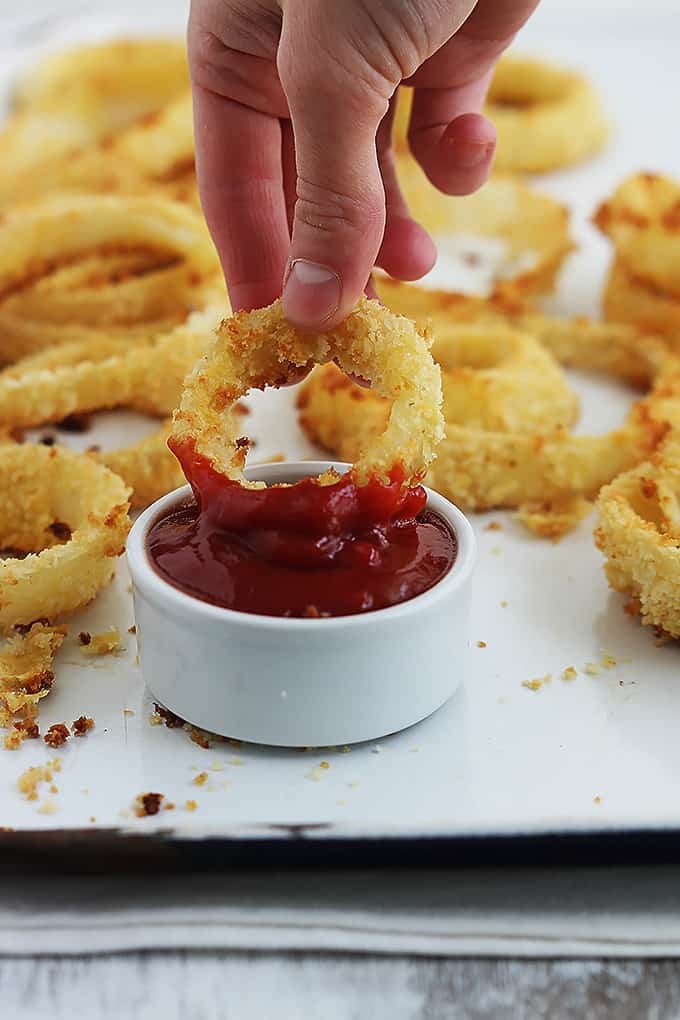 a hand dipping an onion ring in a dipping bowl of ketchup with more onion rings on the side.