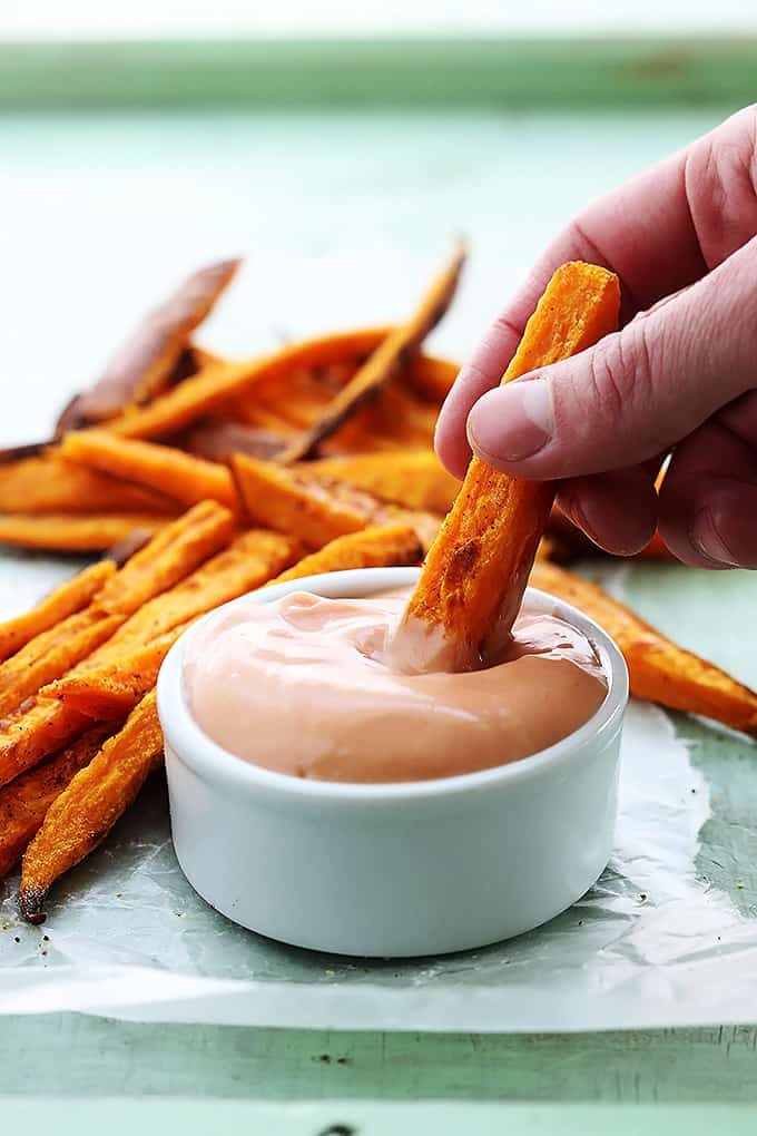 a hand dipping a sweet potato fry in a bowl of fry sauce with more fries in the background.