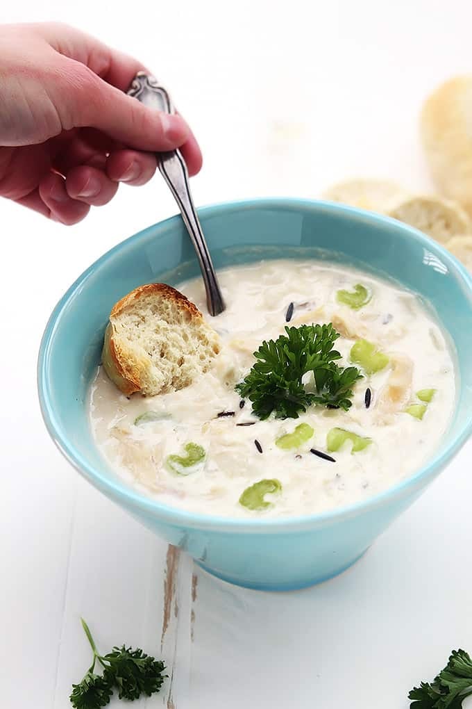 a hand holding a spoon inside a bowl of chicken & wild rice soup with a slice of bread also dipped in the soup.