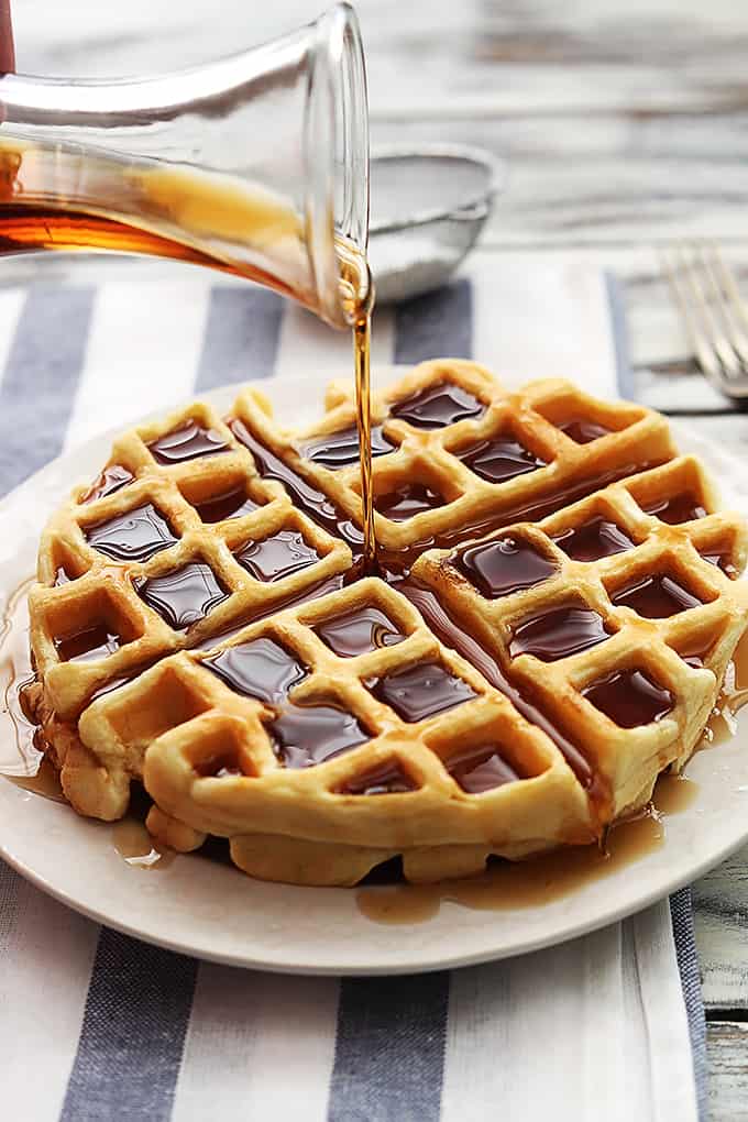 a Dutch cream waffle with syrup being poured on top with a fork and a sifter filled with powdered sugar in the background.