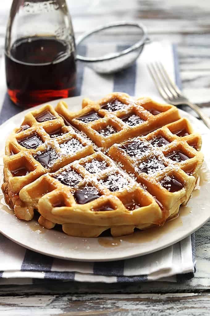 a Dutch cream waffle on a plate topped with syrup and powdered sugar with a jar of syrup, a sifter with powdered sugar, and a fork in the background.