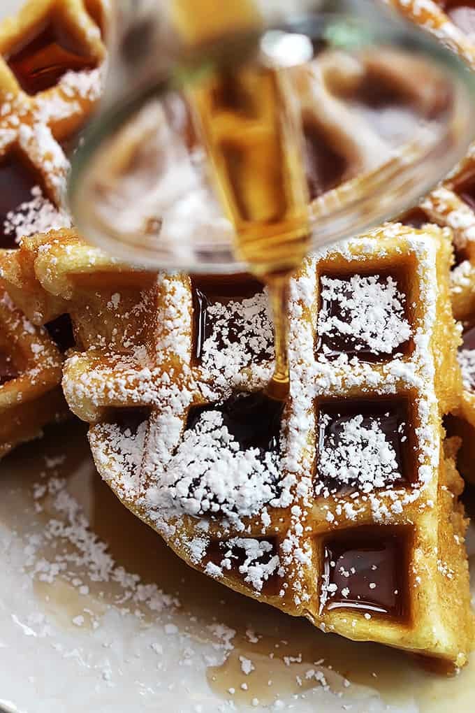 close up of a Dutch cream waffle topped with syrup and powdered sugar with more syrup being poured on top.