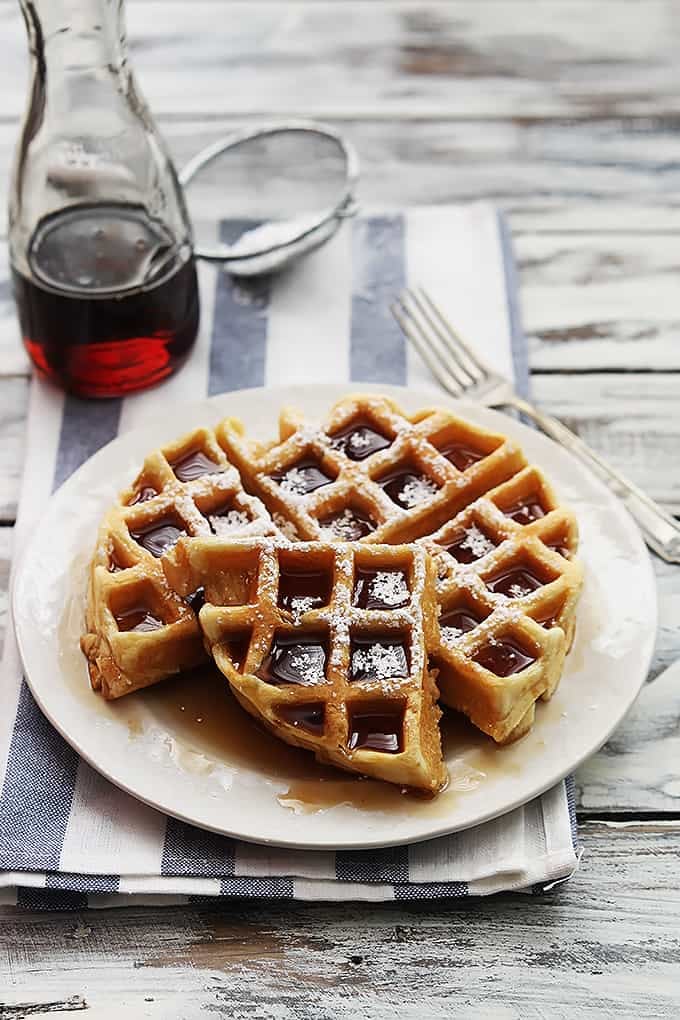 a Dutch cream waffle topped with syrup and powdered sugar on a plate with a jar of syrup, a sifter with powdered sugar and a fork in the background.