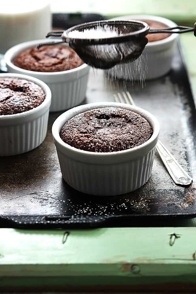 a sifter sifting powdered sugar on top of a flourless chocolate almond cake in a small bowl with more bowls of cake and a fork in the background.