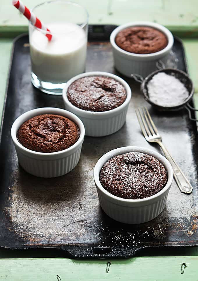 flourless chocolate almond cakes in small individual bowls, a fork, a sifter full of powdered sugar, and a glass of milk on a baking sheet.