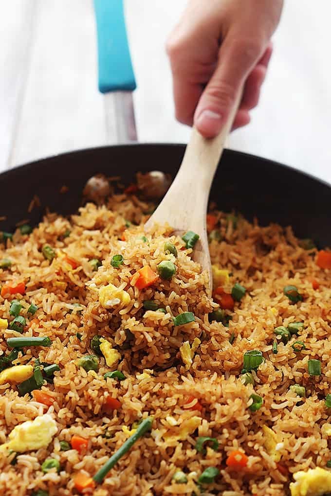 a hand scooping a spoonful of fried rice from a skillet with a wooden serving spoon.