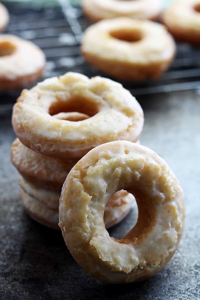 stacked old fashioned sour cream donuts with another donut leaning on the front of them with more donuts on a cooling rack in the background.