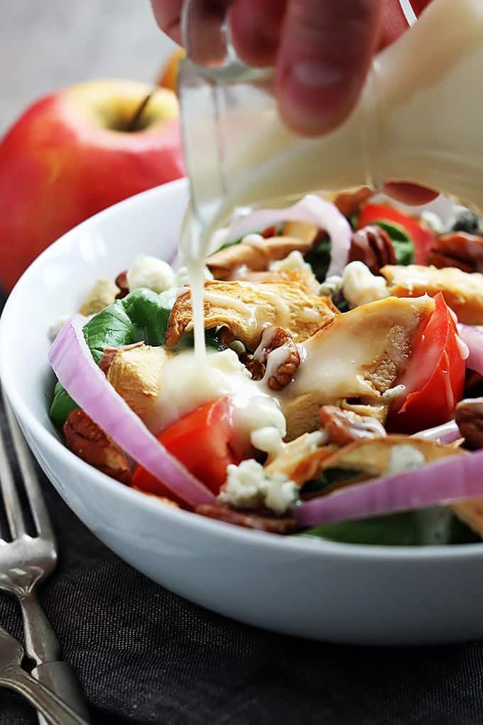 a hand pouring dressing on top of Panera Bread's Fuji apple chicken salad in a bowl with forks and apples on the side.