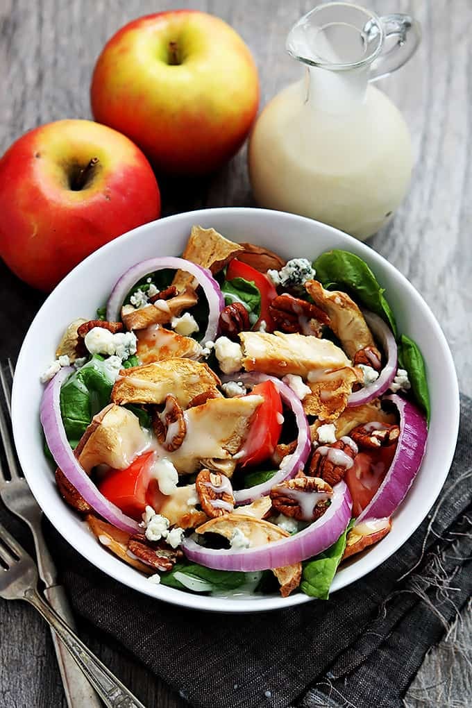 Panera Bread's Fuji apple chicken salad in a bowl with forks, apples, and a glass jar of dressing on the side.