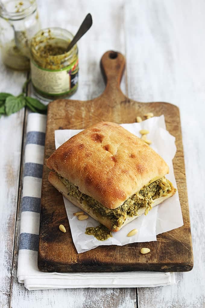 a pesto chicken sandwich on a wooden cutting board with jars of pesto on the side.