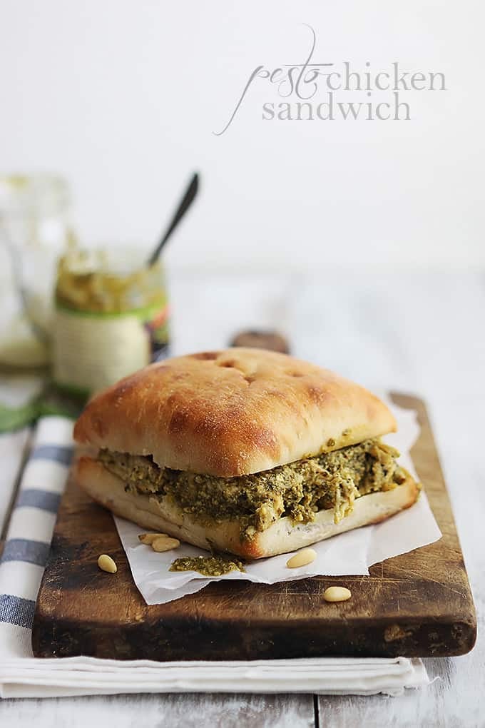 a pesto chicken sandwich on a wooden cutting board with jars pesto in the background with the title of the recipe written on the top right corner of the image.