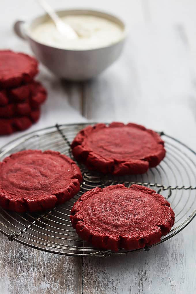 red velvet sugar cookies on an iron rack with more cookies stacked in the background next to a bowl of frosting.