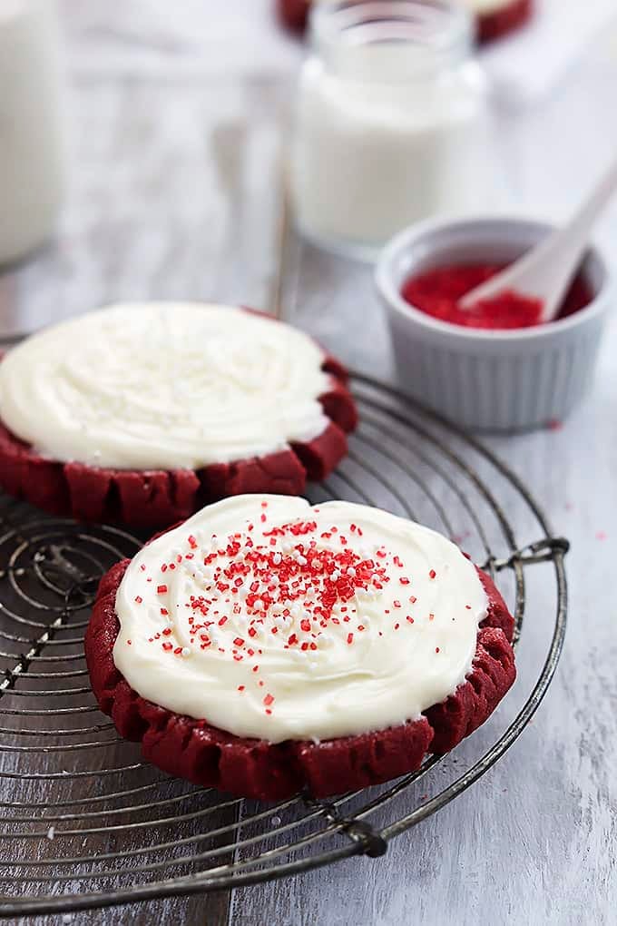 frosted red velvet sugar cookies on an iron rack with the front cookie having sprinkles on it with a bowl of sprinkles in the background.