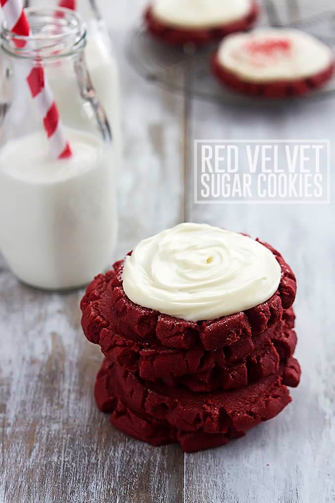 stacked red velvet sugar cookies with the top cookie being frosted next to a jar of milk with the title of the recipe written on the right side of the image.