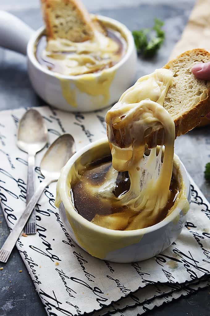 a hand lifting a slice of bread just dipped in a bowl of French onion soup with another bowl of soup with a slice of bread in the background and spoons on the side.