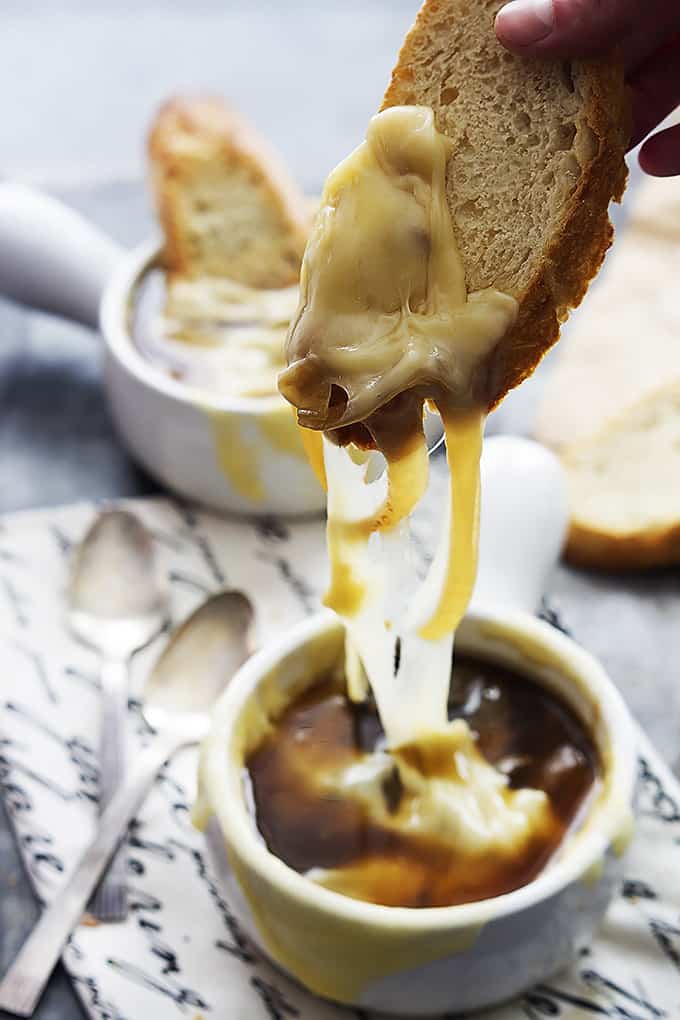 a hand lifting a slice of bread just dipped in a bowl of French onion soup with another bowl of soup with a slice of bread in the background and spoons on the side.