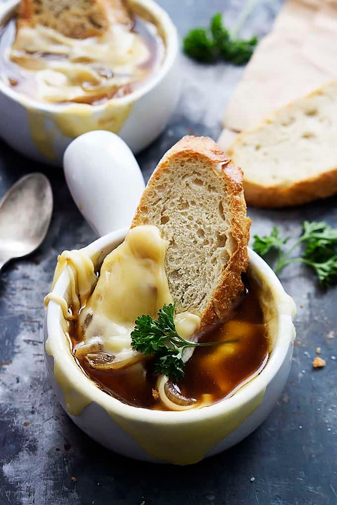 French onion soup with a slice of bread in a bowl with another bowl of soup, a spoon and more bread in the background.