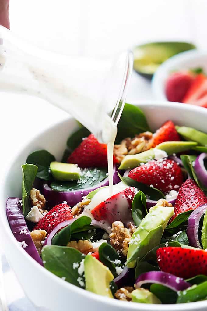 dressing being poured on top of strawberry avocado spinach salad in a bowl.