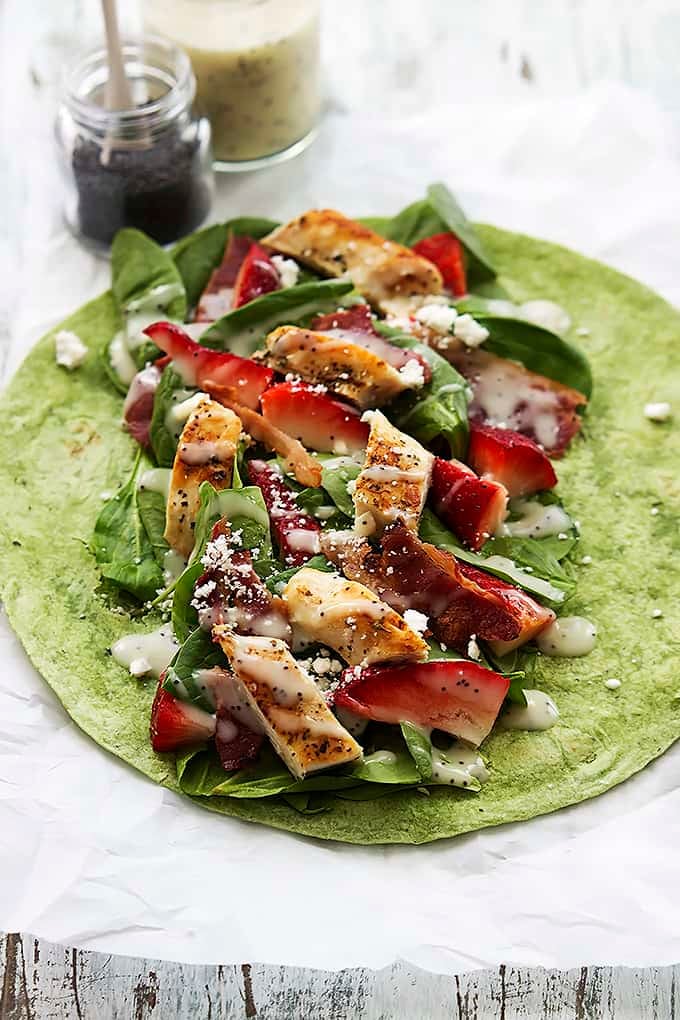 a strawberry chicken bacon & spinach wrap with poppyseed dressing open on a table with a jar of dressing and a jar of poppyseeds on the side.