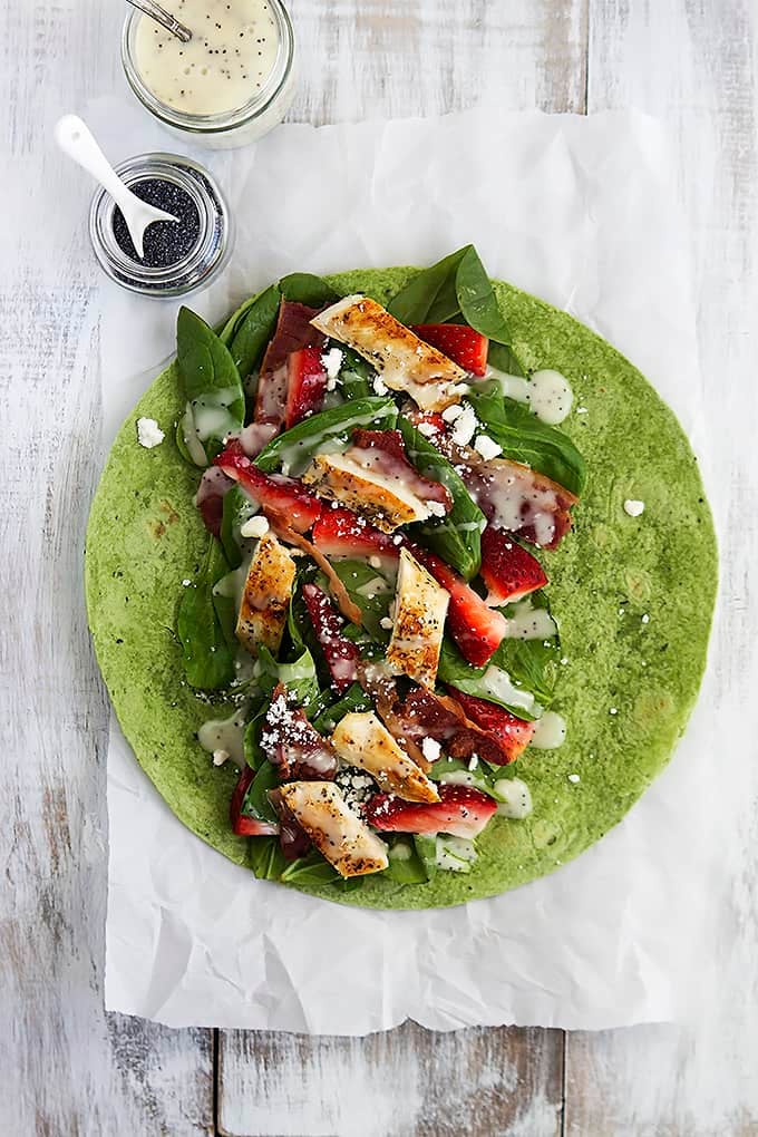 top view of a strawberry chicken bacon & spinach wrap with poppyseed dressing open on a table with a jar of dressing and a jar of poppyseeds on the side.