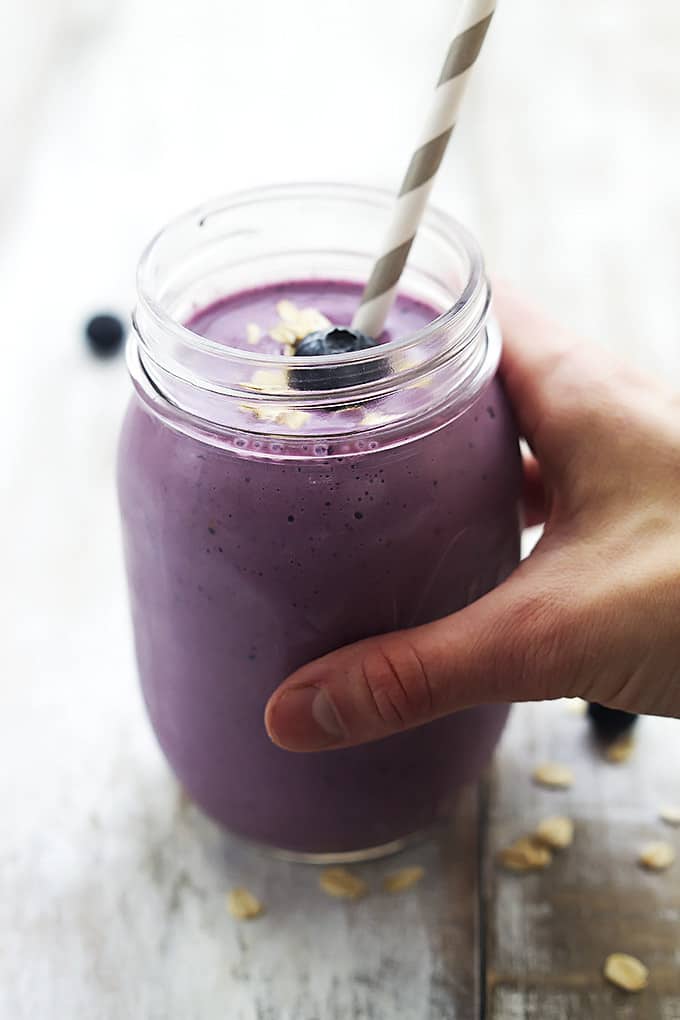 a hand holding a berry oat breakfast smoothie in a jar with a straw and topped with oats and a blueberry.