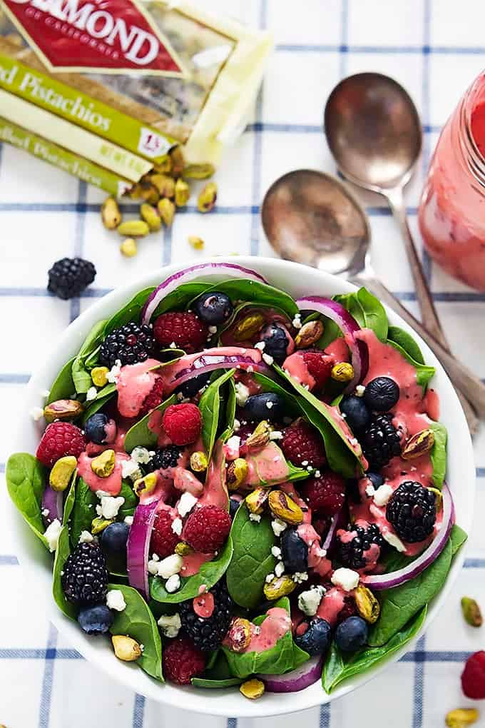 top view of berry lover's spinach salad with berry vinaigrette in a bowl with spoons, a jar of vinaigrette, and a bag of almonds on the side.
