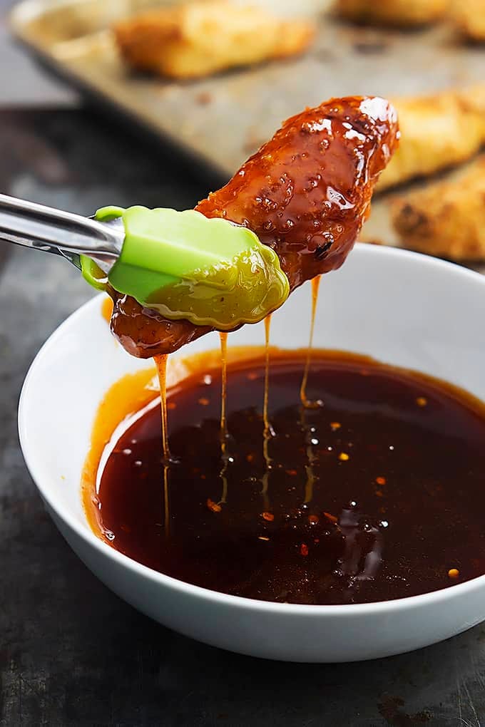 a pair of tongs holding a baked firecracker chicken tender just dipped in sauce above a bowl of sauce.