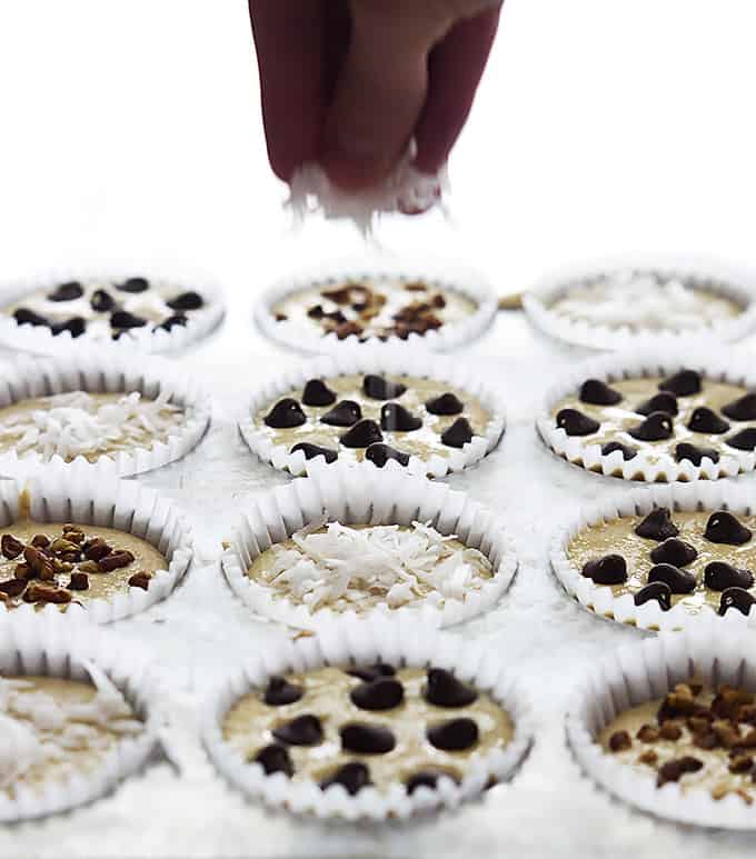 unbaked flourless banana blender muffins in a muffin tin with a hand sprinkling coconut shavings on top of one with the others having chocolate chips, nuts, or coconut shavings on top.