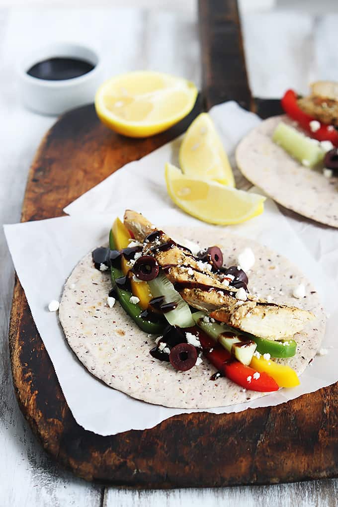 a Greek chicken taco laid flat with a lemon half and slices with another taco all on a wooden cutting board with a small bowl of sauce on the side.