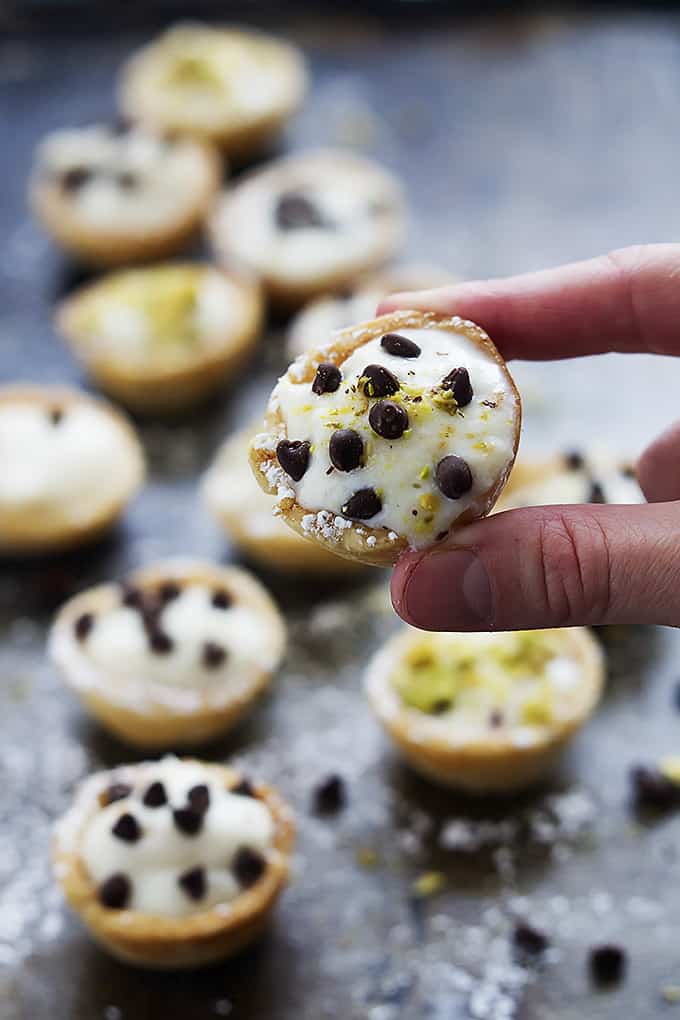 a hand holding up a cannoli bite with powdered sugar, chocolate chips and pistachios on top with more bite in the background.
