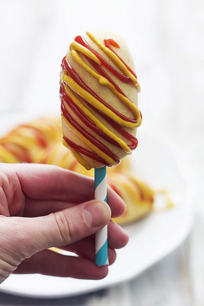 a hand holding up a homemade baked corn dogs on a stick with mustard and ketchup on it.