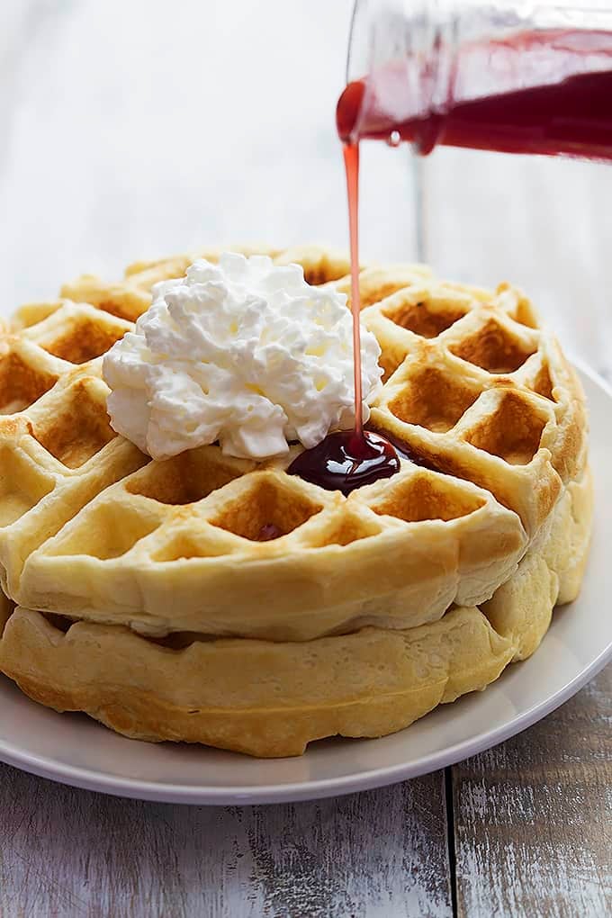 Greek yogurt waffles topped with whipped cream on a plate with strawberry syrup being poured on top.