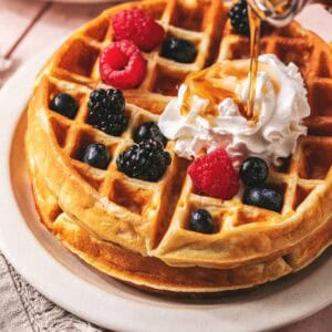 Syrup being poured onto a greek yogurt waffles with berries and cream on a plate.