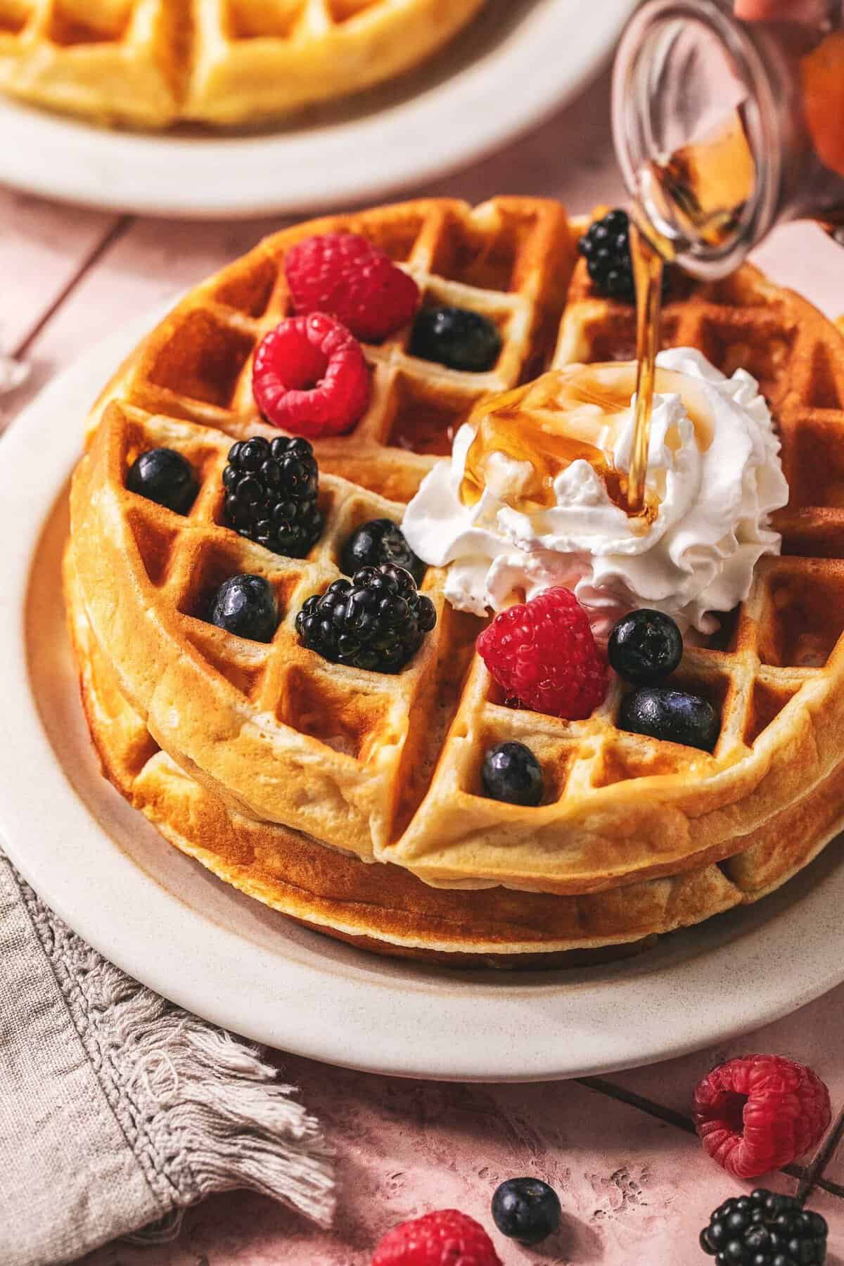 Syrup being poured onto a greek yogurt waffles with berries and cream on a plate.
