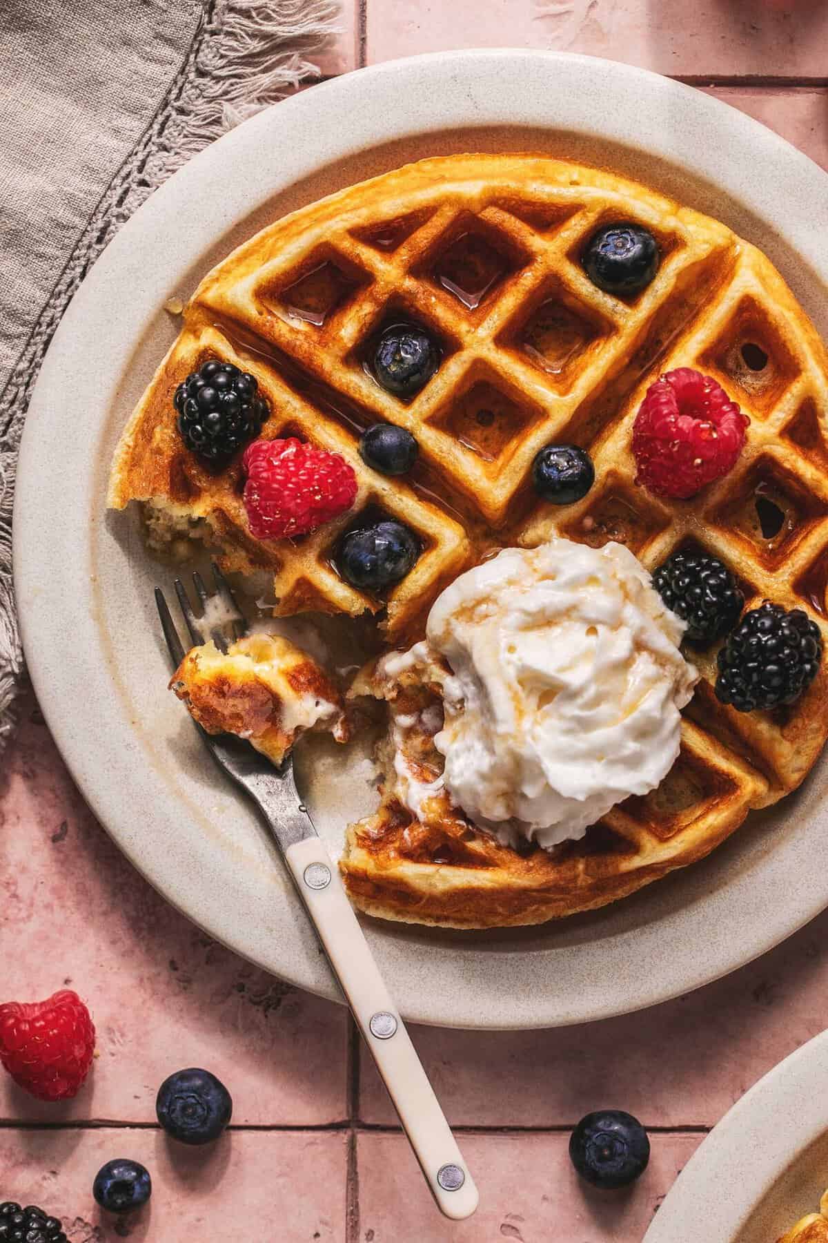 Overhead of a greek yogurt waffles with berries and cream and a fork with a bite on it on a plate.