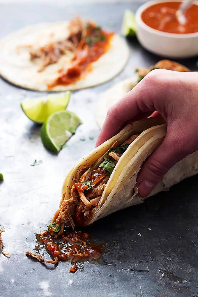 a hand picking up a slow cooker pork carnita with another carnita, slices of lime, and a bowl of sauce in the background.