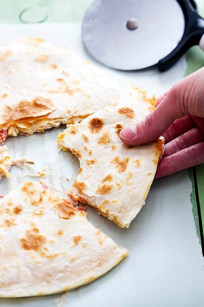 a hand picking up a slice of a slow cooker chicken quesadilla with a bite missing with more quesadilla slices and a pizza cutter on the side.