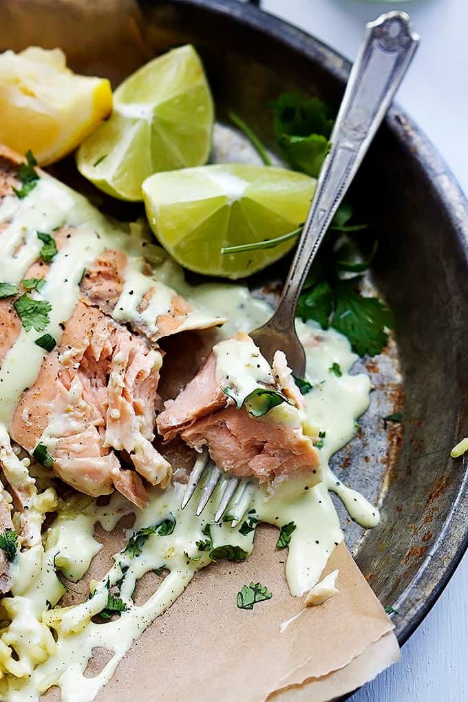 a bite of baked salmon with avocado cream sauce on a fork with more salmon and slices of limes on the side on a plate.