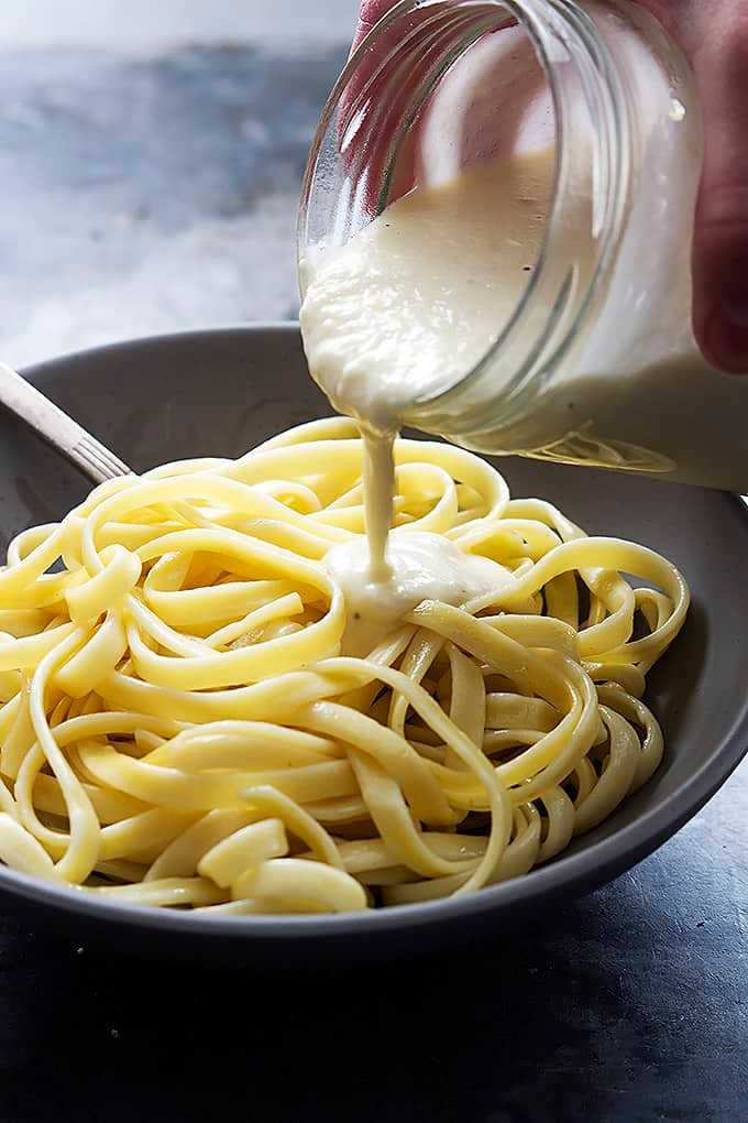 a jar of Greek yogurt alfredo sauce being poured on top of fettucine noodles in a bowl.