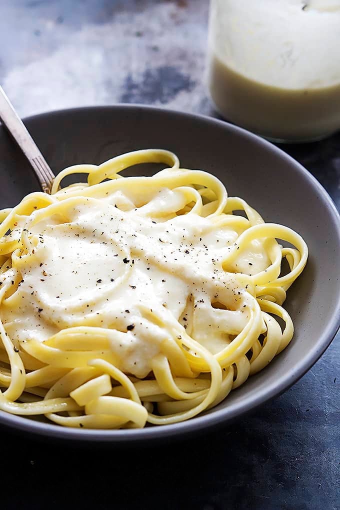 fettucine noodles with Greek yogurt alfredo sauce on top with a fork in a bowl with a jar with more sauce on the side.