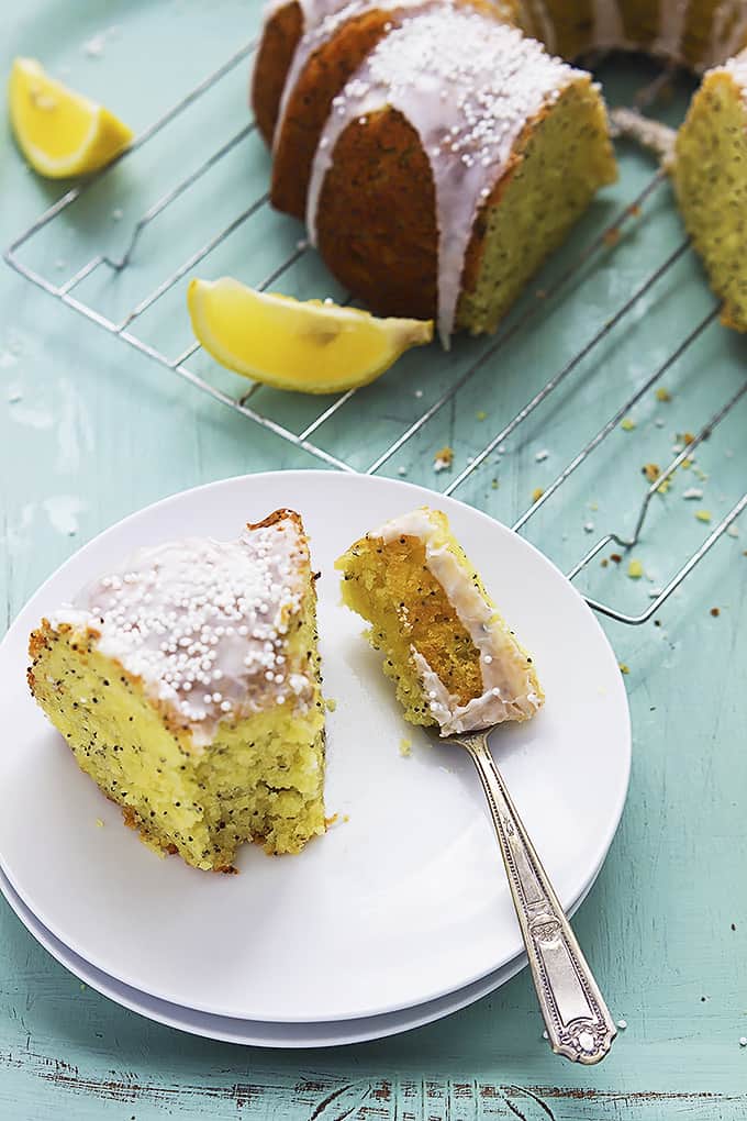 a piece of lemon poppyseed bundt cake with a bite on a fork on a plate with the rest of the cake and lemon slices in the background.