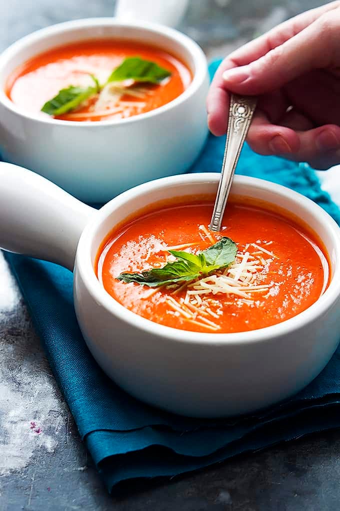 a hand holding a spoon in a bowl of roasted red pepper soup with another bowl of soup in the background.