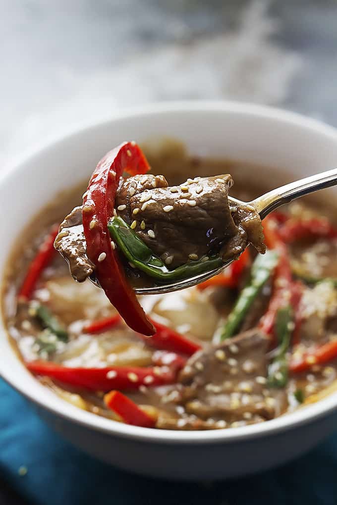 a spoon being held up with slow cooker sesame beef on it with a bowl of more beef in the background.