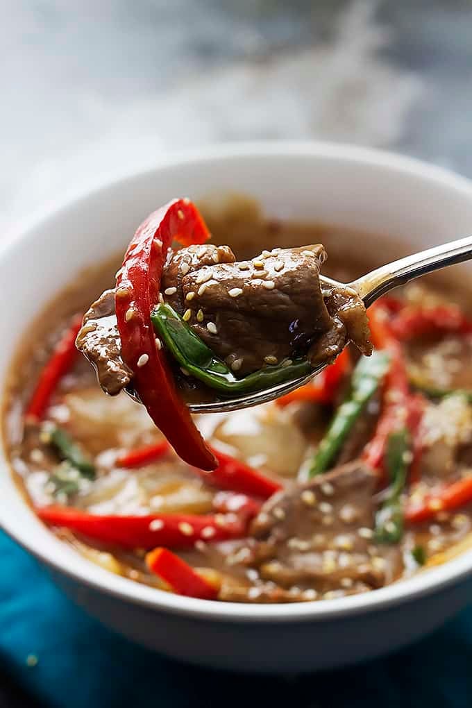 a spoon being held up with slow cooker sesame beef on it with a bowl of more beef in the background.