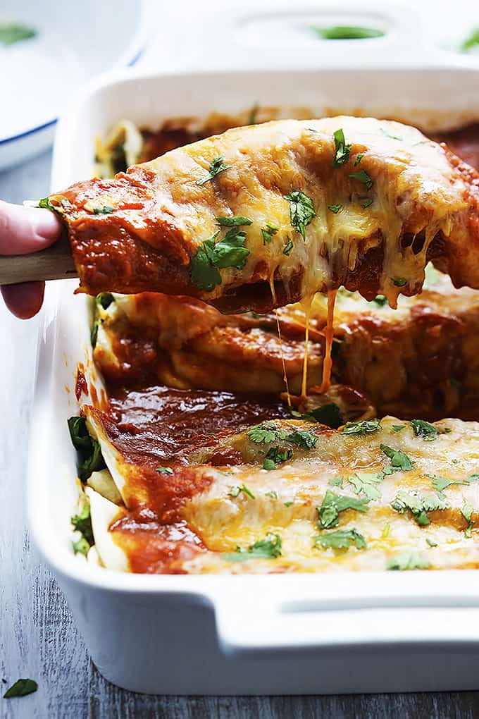 a hand scooping a cheesy spinach enchilada with a spatula from a cooking pan of enchiladas.