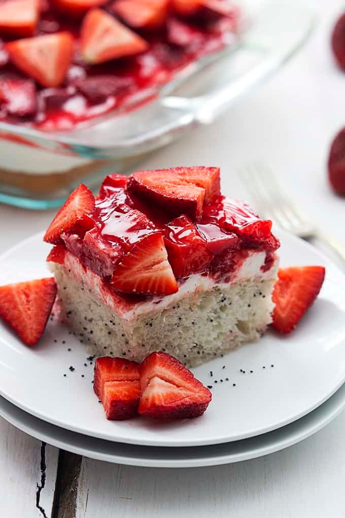 a piece of strawberry poppyseed cake on a plate with more cake in a glass pan in the background.