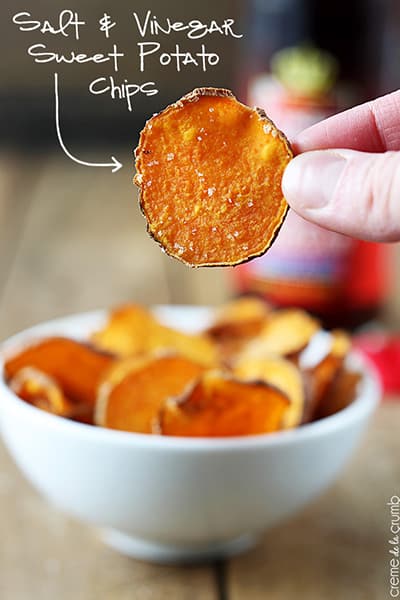 a hand holding up a baked salt & vinegar sweet potato chip with a bowl of chips in the background with the title of the recipe with an arrow pointing to the chip written on the top left corner of the image.