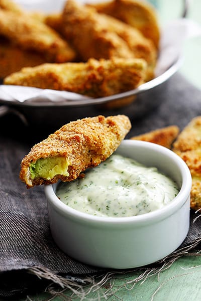 a baked avocado fry sitting on the side of a bowl of dipping sauce with more fries in a bowl in the background.