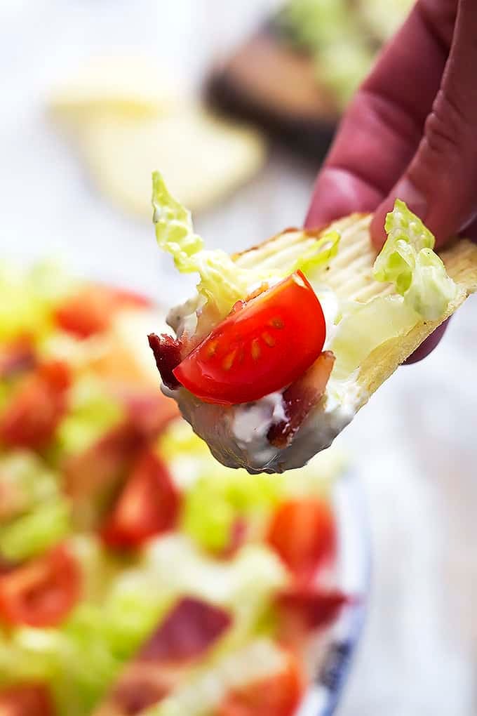 a hand holding up a chip dipped in creamy Ranch BLT dip above a bowl of dip.