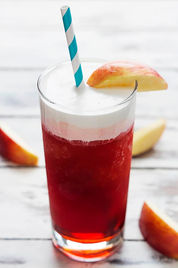 a glass of Red's apple slush (Disneyland copycat) with a slice of an apple on the top of the glass along with a straw and other slices of apple around it.